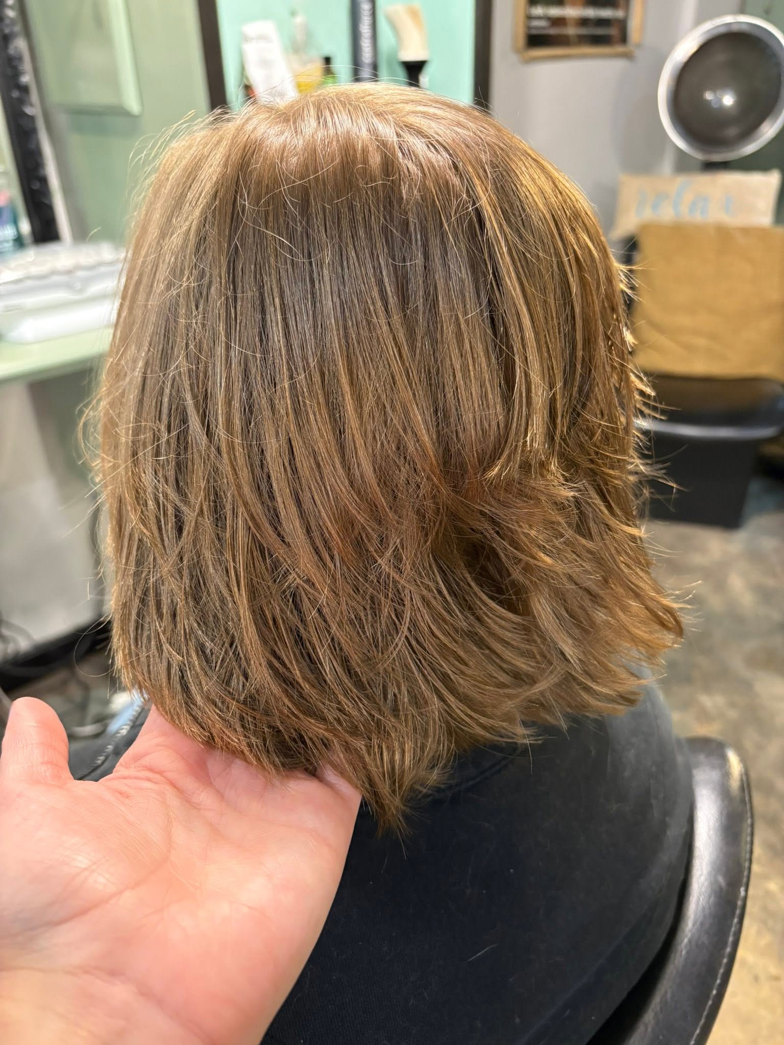 A hand lifts the layered ends of a fresh, shoulder-length chestnut haircut in a salon, showing soft texture and warm highlights; polished, relaxed mood.