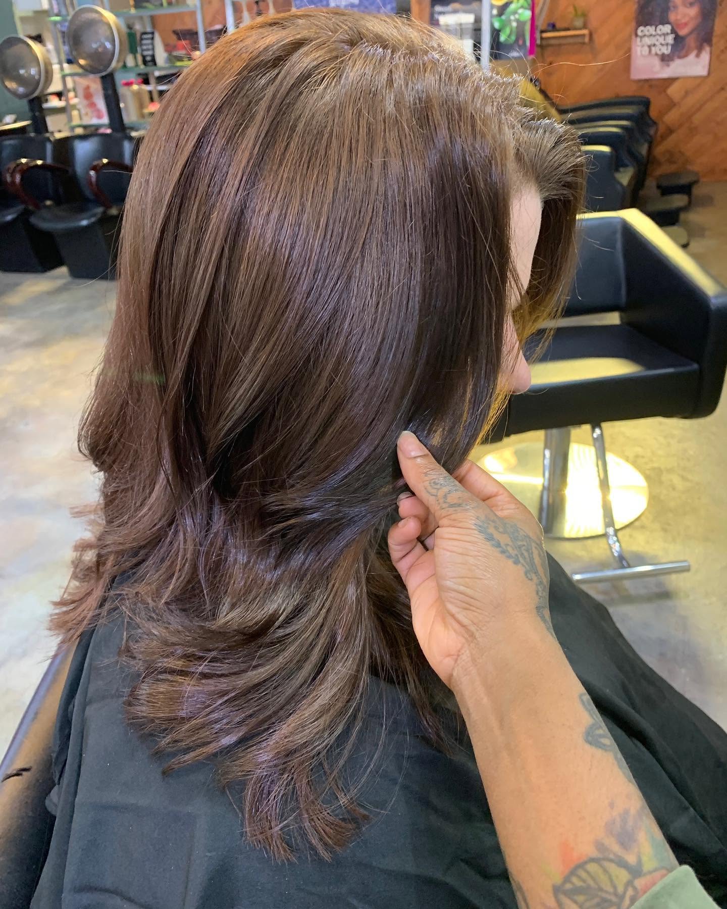A stylist lifts the softly layered ends of a client’s glossy, chestnut-brown hair in a salon, showcasing the smooth, voluminous cut.