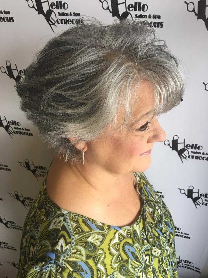 A woman in profile shows off a voluminous silver-gray layered hairstyle, wearing a patterned green blouse and dangling earrings against a salon-branded backdrop.