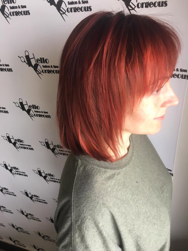A woman with a sleek, chin-length auburn bob and side-swept bangs poses in profile against a salon-branded backdrop, showing off the polished haircut.
