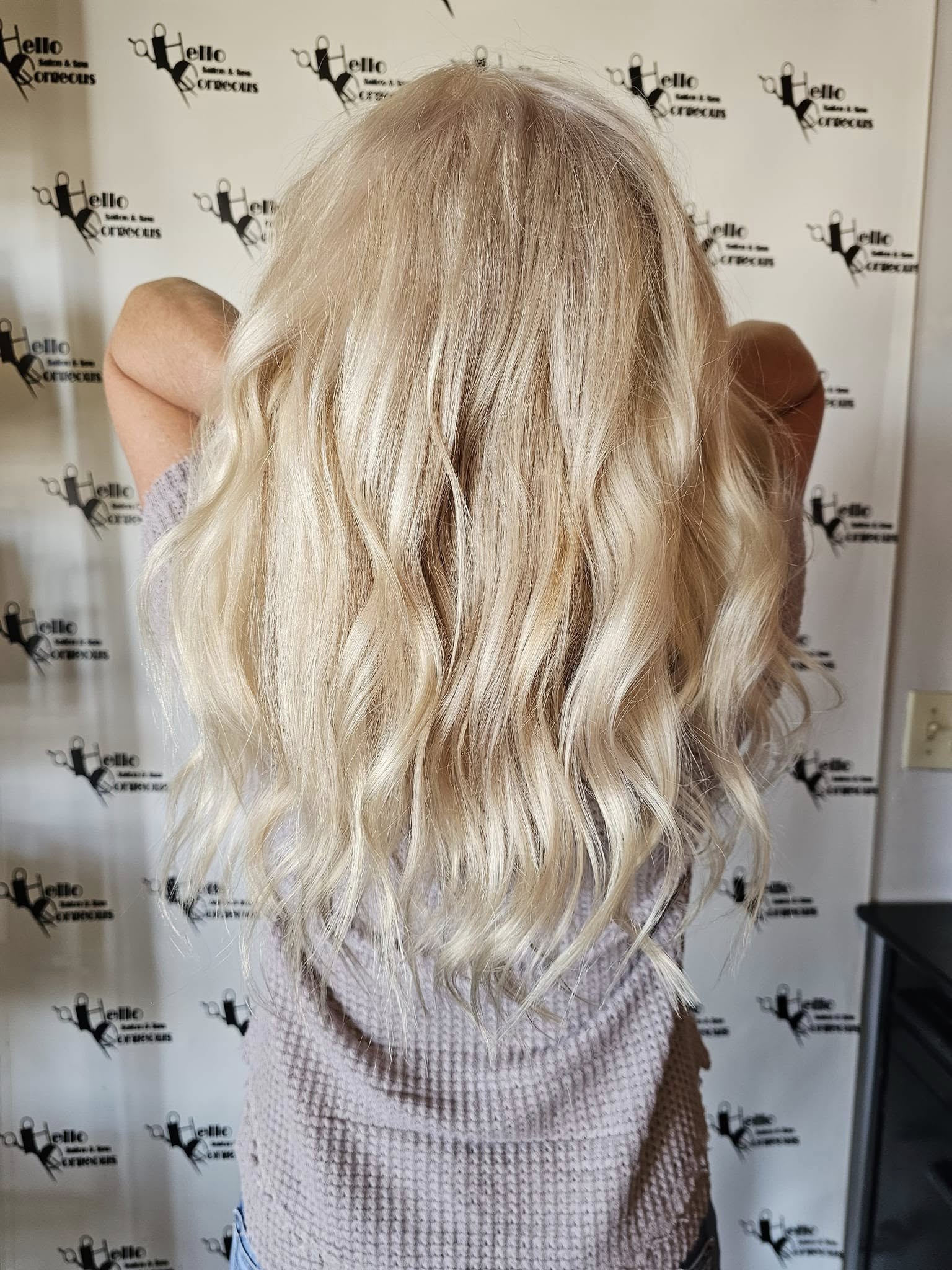 Back view of a woman showing long, blonde, softly wavy hair at a salon event, with her arms raised against a branded photo backdrop. The mood is polished and relaxed.