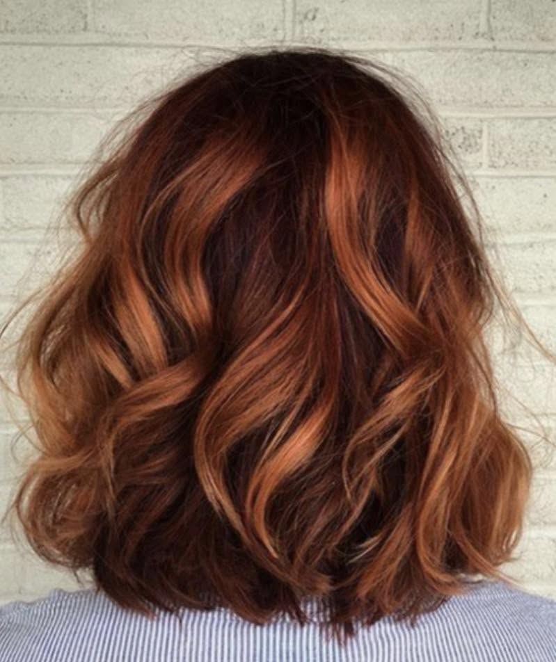 Back view of a woman’s shoulder-length wavy auburn hair with rich copper highlights and layered texture, styled in a tousled bob against a pale brick wall.