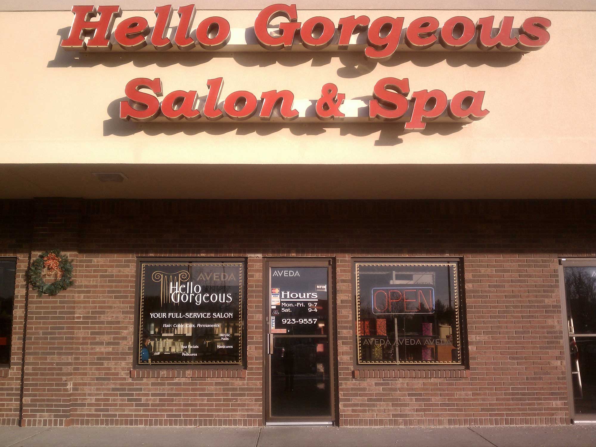 Red lettering announces “Hello Gorgeous Salon & Spa” above a brick storefront, with salon windows, an hours sign, and an “OPEN” sign in the window.